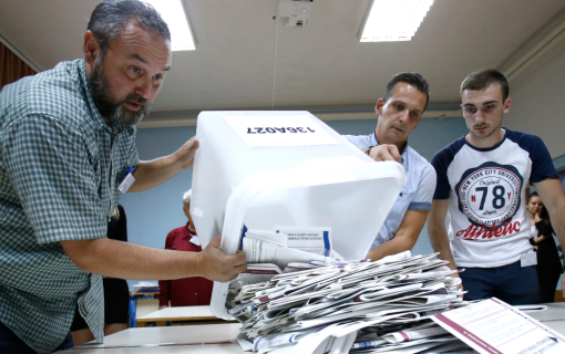 Three Bosnian election workers empty paper ballots from a container onto a table.