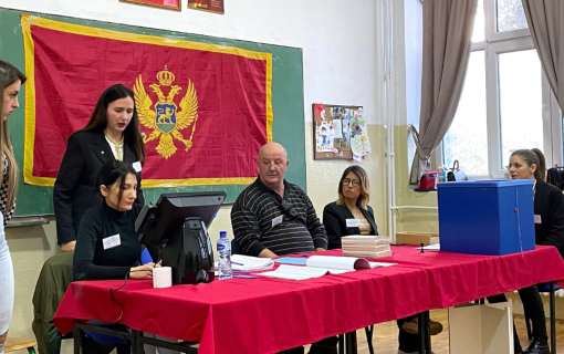 Four people sit at a red table, while two people stand behind the table. A Montenegro flag is hanging up on the wall behind them.