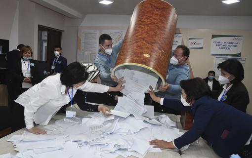 Several people wearing masks stand around a table, pulling pieces of paper out from a large, upside-down container held up by two men.