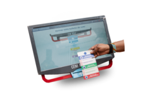 Image of a ballot voting machine that will be utilized in the 2023 DRC elections. There is a hand holding a ballot to be scanned in front of the machine.