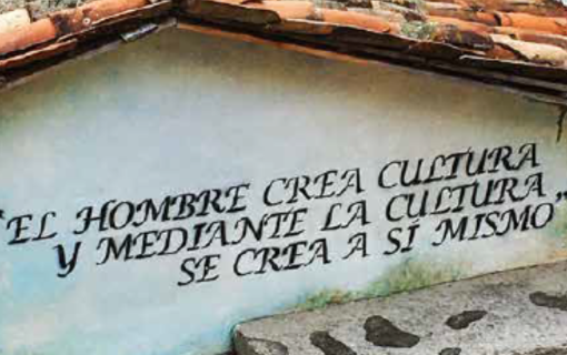 A weathered building wall with a Spanish quote painted on it, &quot;El hombre crea cultura y mediante la cultura se crea a sí mismo,&quot; under a rustic tiled roof.