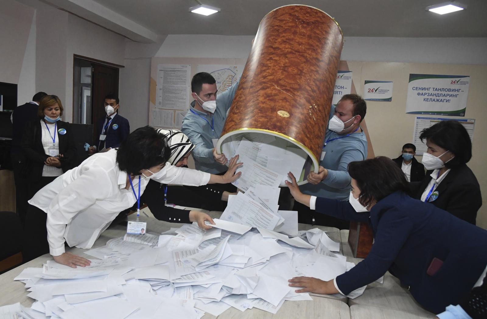 Several people wearing masks stand around a table, pulling pieces of paper out from a large, upside-down container held up by two men.