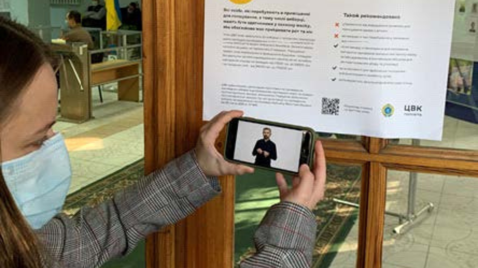 A woman holds up a smartphone to scan a QR code on a poster. Her smartphone plays a video of a person using sign language.