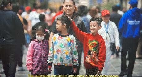 Family walking through crowded streets. Yellow text reads "The organizing principle behind democracy is helping people"