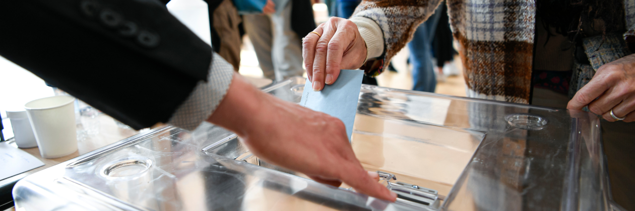 picture shows a person voting with a ballot paper in its envelope just before being placed in the ballot box