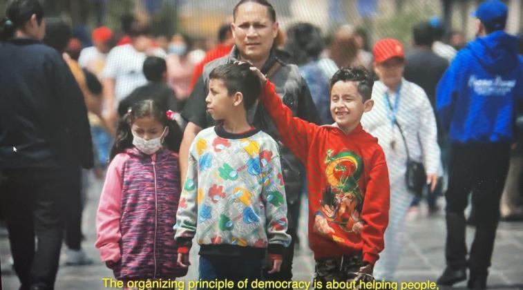 Family walking through crowded streets. Yellow text reads "The organizing principle behind democracy is helping people"