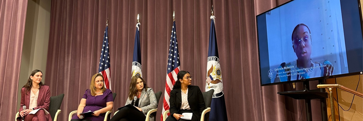 Gabrielle McKenzie on tv screen at USG event. Panelists seated in person to her left in front of flags.