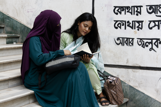 young women sit on steps one in a niquab one uncovered reading a book together. 