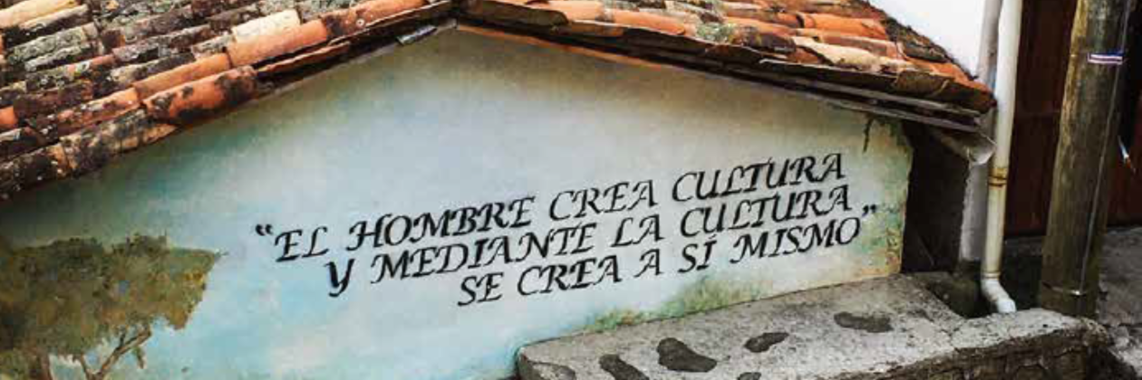 A weathered building wall with a Spanish quote painted on it, "El hombre crea cultura y mediante la cultura se crea a sí mismo," under a rustic tiled roof.