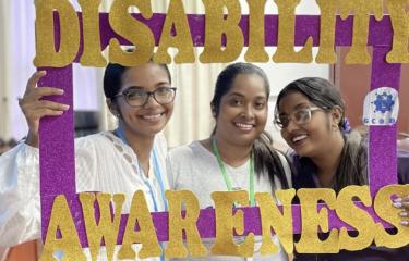 Alt text: Three women from Guyana smile at the camera. They are holding up a purple photo frame that reads “Disability Awareness” in gold letters.