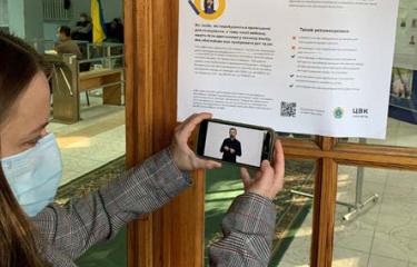 A woman holds up a smartphone to scan a QR code on a poster. Her smartphone plays a video of a person using sign language.