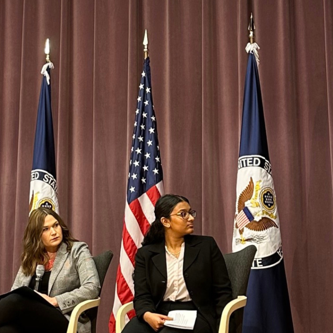 Gabrielle McKenzie on tv screen at USG event. Panelists seated in person to her left in front of flags.
