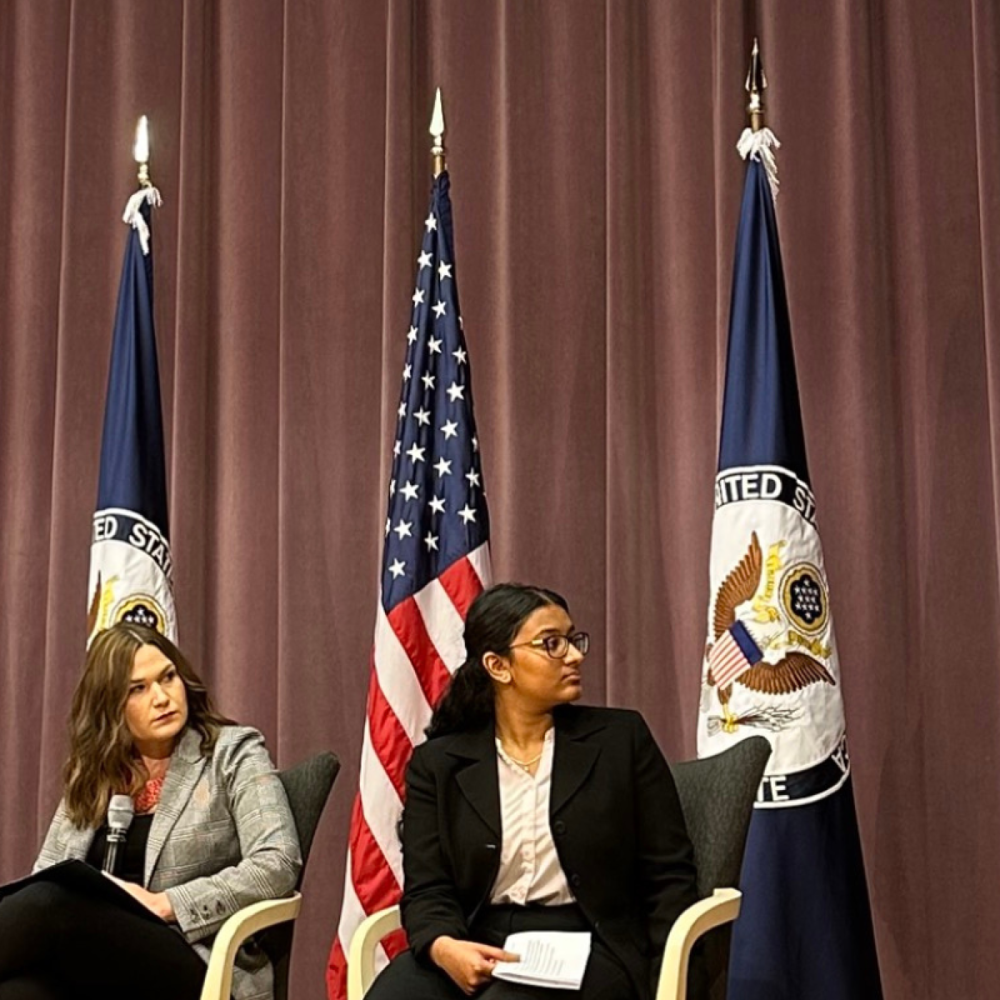 Gabrielle McKenzie  on tv screen at USG event. Panelists seated in person to her left in front of flags. 