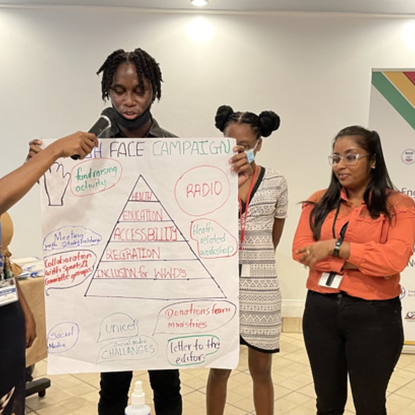 Five young participants of IFES Guyana's ENGAGE program present their work to their peers. A participant is holding the microphone up to another participant holding a poster board as their teammates listen.